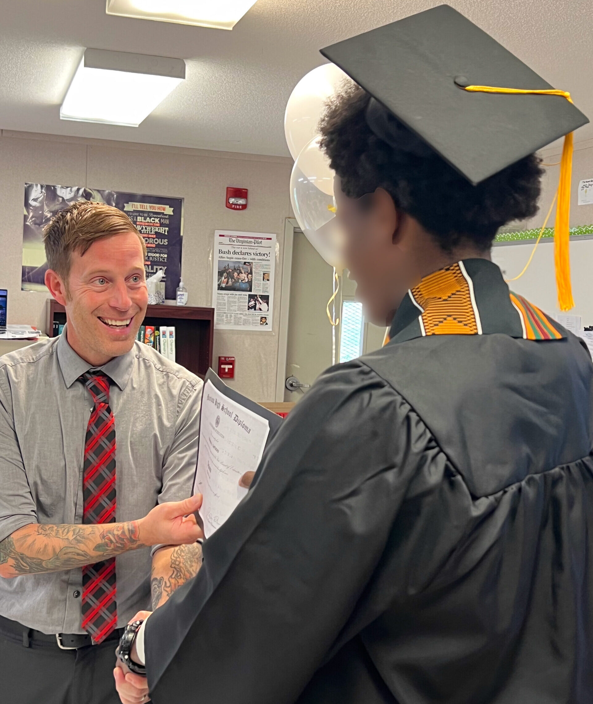 A recent high school graduate wearing a black cap and gown shakes hands with his principal as he receives his diploma.