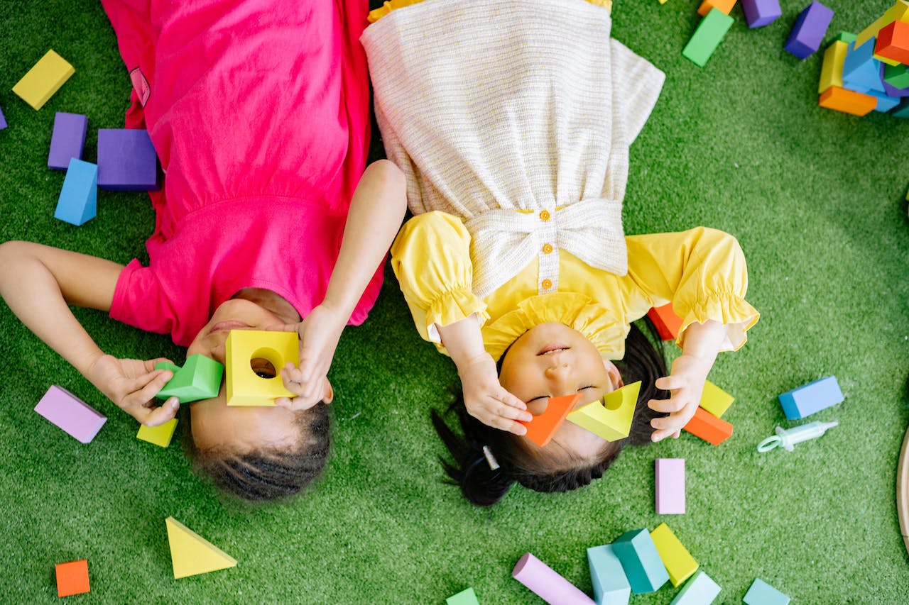 Two young girls lay on a green carpet, playing with colorful blocks.